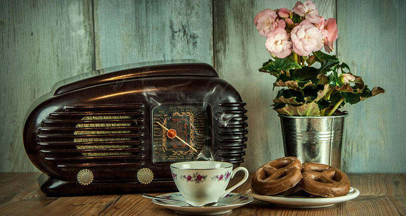En gammal radio, en rosa blomma i en metallkruka, en blommig kaffekopp och chokladkringlor som liggerpå en träytan. En blå trävägg i bakgrunden.