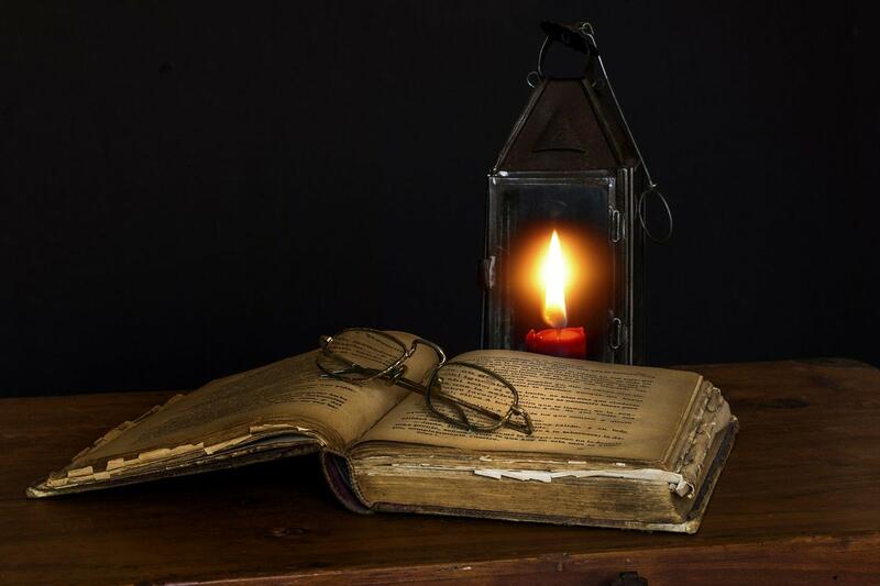 En gammal bok ligger uppslagen på ett bord. Ett par glasögon ligger på boksidorna, en ljuslykta med ett rött ljus lyser i bakgrunden.
